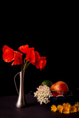 Still life on a black background. A bouquet of wild red poppies in a flacon. Bowl with fruits - pomegranate, apple and avocado. Copy space.