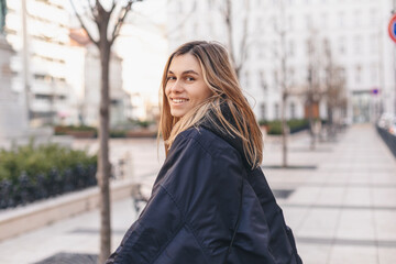 Fototapeta premium Happy blonde woman wear navy blue bomber jacket walking in the street and smiling to camera. Stylish girl in fashion outfit walking over the city. What a beautiful day out. Girl turn around.