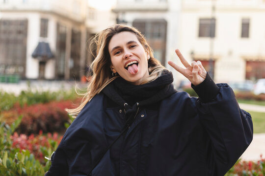 Cool blonde woman in navy blue bomber shows sign v, peace and friendship with two hands, show tongue and posing outside. Happy woman walk and posing outdoor in the city.