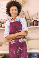 A cheerful and attractive African American woman stands in a studio, her arms crossed as she smiles for the camera. She is an artist, creating beautiful traditional artwork with paint and sketching.