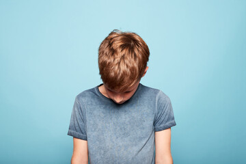 Unhappy young caucasian boy looking down, isolated against a blue background.