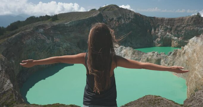 Tourist woman rises hands over volcanic Kelimutu blue crater lakes. Nature background. Travel destination. Beautiful wild landscape. Exotic summer vacation, hiking, relax. Close up back view 4K