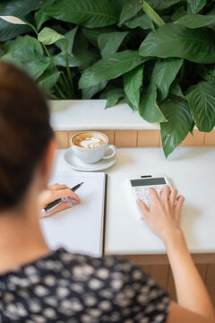 Above top close up back view working woman with note papers and calculator calculating financial. Women seller online note order on paper and calculate sales with favorite cup of coffee hot latte