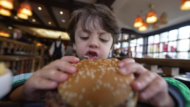Hungry small boy eating burger sitting inside restaurant place. One male caucasian child takes a bite of hamburger. Kid eating delicious food