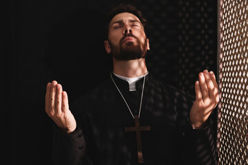 Catholic priest in cassock praying to God in confessional booth - obrazy, fototapety, plakaty