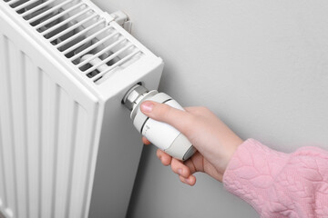 Girl adjusting heating radiator thermostat near white wall indoors, closeup