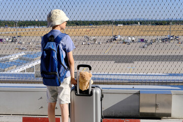 child in outdoor terrace of airport, young traveler is waiting for boarding, boy of 8-10 years old in blue shirt with suitcase and teddy bear, concept of vacation, long journey, air travel insurance