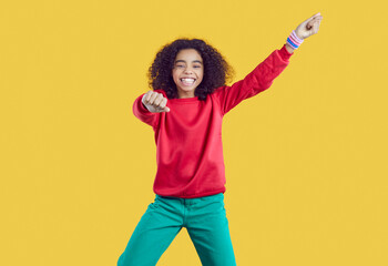 Happy child having fun in studio. Portrait of funny kid in casual outfit dancing isolated on yellow background. Pretty African American girl in red sweat shirt and green jeans dancing gangnam style