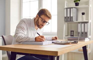 Busy financial accountant working at his office desk. Young man in a white shirt and glasses sitting at his office table, doing business paperwork, and using a modern laptop