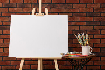 Wooden easel with blank canvas and different art supplies near brick wall, closeup