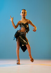 Expressive dance. Beautiful little girl in stage attire dancing latino madern ballroom dance over blue background. Music, dance, education, professional dance school concept