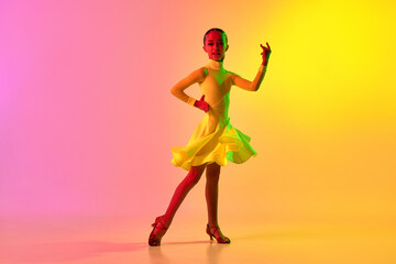 Fototapeta na wymiar Emotional little girl in yellow stage dress dancing classical ballroom dance over gradient pink-yellow background in neon light filter.