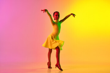 Emotional little girl in yellow stage dress dancing classical ballroom dance over gradient pink-yellow background in neon light filter.