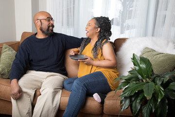 Life style portrait of a black married couple sitting on the couch and enjoying each other's company. The woman is eating an oatmeal muffin. 