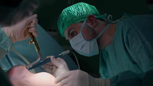 Side view focused Middle Eastern surgeon using forceps in cut breast of patient on surgical table. Concentrated expert doctor performing surgery in operating room. Bust augmentation transplantation