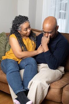 Portrait Of A Middle Aged Black Married Couple Sitting On The Couch. The Wife Is Comforting Her Husband While He Is Distressed. 