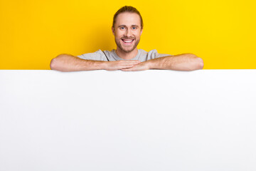 Photo of optimistic man with long hairstyle wear gray t-shirt lean hands on white placard empty space isolated on yellow color background