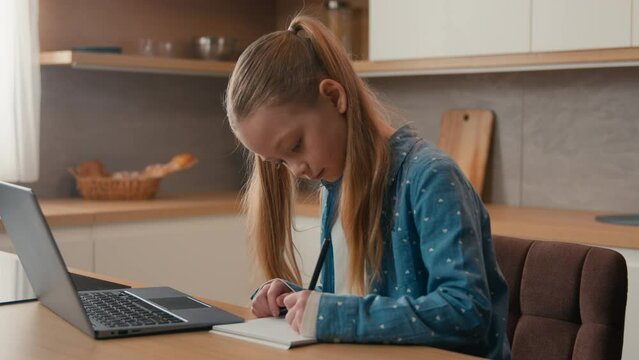 Cute primary Caucasian school girl studying at kitchen with laptop. Child daughter kid schoolgirl pupil doing homework writing exercise distance learning. Children home education e-learning quarantine