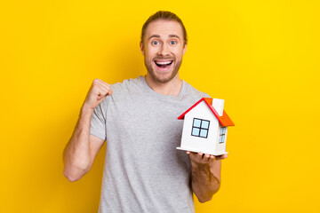 Photo of positive lucky man wear grey t-shirt holding small house rising fist isolated yellow color background