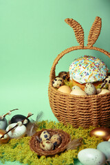 Easter decorations in a basket with rabbit ears. Easter cake and eggs on moss, quail eggs and feathers on mint green background. pussy willow Place for text.