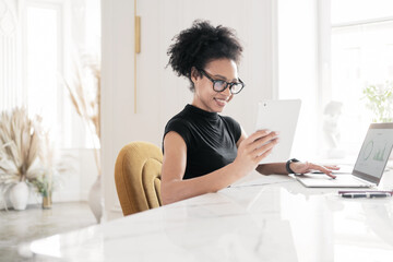A young woman with glasses working at a workplace in the office, using a computer and tablet. Website designer online project.