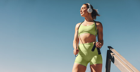 Female athlete in her 30s outdoors taking a break from her cardio workout, holding a skipping rope and wearing sportswear - obrazy, fototapety, plakaty