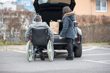 A woman helps a disabled person on a wheelchair to pack groceries into the car