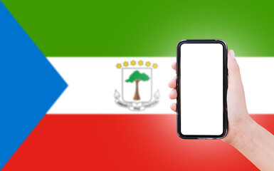 Male hand holding smartphone with blank on screen, on background of blurred flag of Equatorial Guinea. Close-up view.