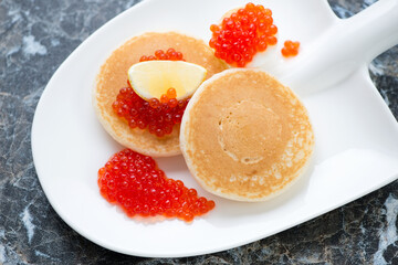Shovel-shaped plate with red caviar and mini pancakes, middle closeup, horizontal shot on a black marble background