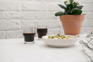 Cottage cheese with fruits and honey in a white plate with a spoon and a napkin, two glasses of coffee and pot with flowers on a white background. Healthy breakfast.