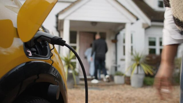 Close up of woman plugging electric charger into car with multi ethnic family walking into home