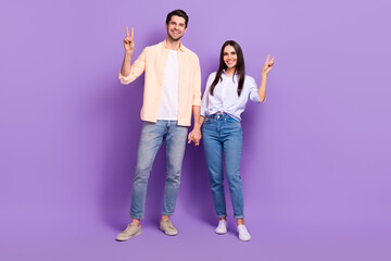 Full body portrait of two cheerful partners hold arms demonstrate v-sign isolated on purple color background