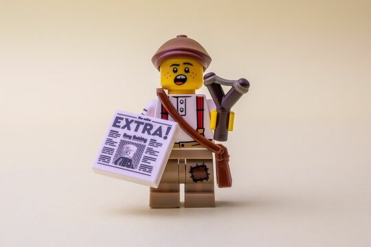 LEGO minifigure toy holding newspaper and slingshot on pastel background