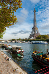 View of the Eiffel Tower with the River Seine at a sunny day in Autumn. House and Restaurantsboats are towed at the River bank. A Couple is enjoying the view.