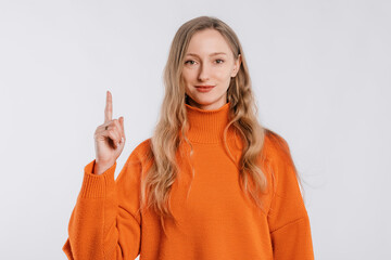 Cheerful attractive young woman points finger up, shows way to banner, demonstrates logo or advertisement on top, stands in an soft orange sweater over neutral background