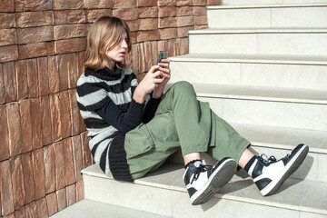 Young pretty woman in a striped black and white sweater, social media influencer, sitting on stairs of the building city using smartphone.