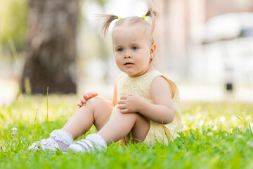 a happy little blonde girl in a yellow dress is sitting on the green grass in the park in summer. Earth Day. Children Protection Day