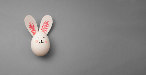 White chicken egg with rabbit paper ears on gray background. Hare muzzle. Easter Bunny. Horizontal banner with space for text. Preparations for celebration of religious holiday Happy Easter. Copyspace