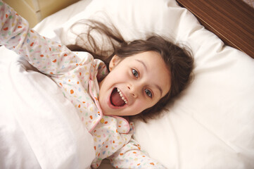 View from above of a mischievous amazed little girl in white pajamas, smiling and making faces looking at camera, stretching while waking up in the morning, relaxing on her comfortable bed