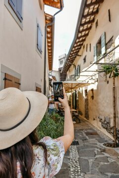 Young woman in a floral sundress and a straw hat taking pictures of the narrow streets of Smartno