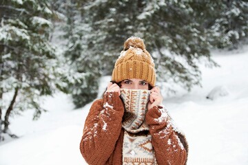 Woman in a cozy winter sweater and hat pulling her scarf over her mouth in a misty winter forest