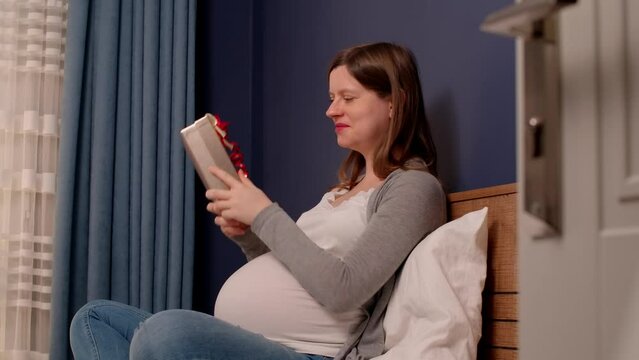 Pregnant woman sitting in bedroom with present box for unborn baby, curious what in the box.