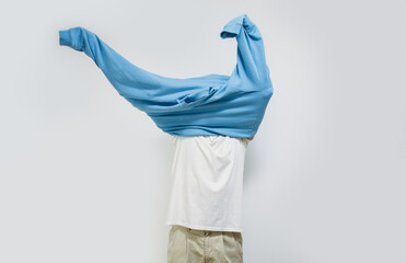 A guy dressed in a white t-shirt and brown trousers is trying on a blue sweatshirt, holding his hands up isolated on a white studio background. The process of putting on a sweatshirt.