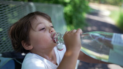 Child drinks water during hot summer day. Little boy gulping from glass bottle hydrating liquid at outdoor park