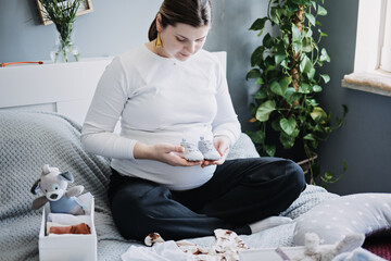Pregnant woman with big belly holding bodysuit and sorting clothes for future. How to Organize...
