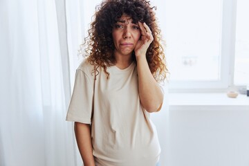 A pregnant woman stands in the house at the window, tired and has a headache, tears, crying from fatigue in a home T-shirt, the complexity of motherhood