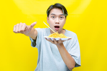 Happy euphoric asian young man in grey tshirt eating instant fried noodle using bamboo chopstick isolated over yellow background. stylish asian man with peek a boo hair eats instant noodle