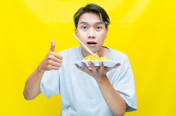 Happy euphoric asian young man in grey tshirt eating instant fried noodle using bamboo chopstick...