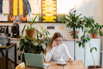 cheerful blogger talking on smartphone near laptop next to cup of cappuccino in cafe.