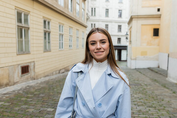 cheerful young woman in blue trench coat standing near buildings on street in Vienna.